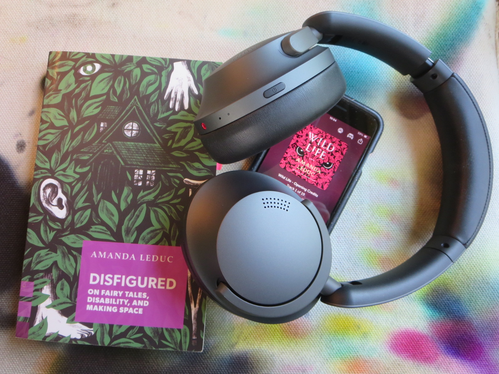 Image of green and purple bookcover, titled "Disfigured: On Fairy Tales, Disability, and Making Space." Headphones and cell phone with pink cover of audio book, titled "Wild Life."