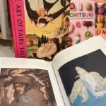 Book cover with image of small woman riding the back of a brown and white bird with black text running vertically along the wide bright pink spine Art of FairyTales. Open pages of the book in front with a picture of a woman wearing a long white dress surrounded by polar bears
