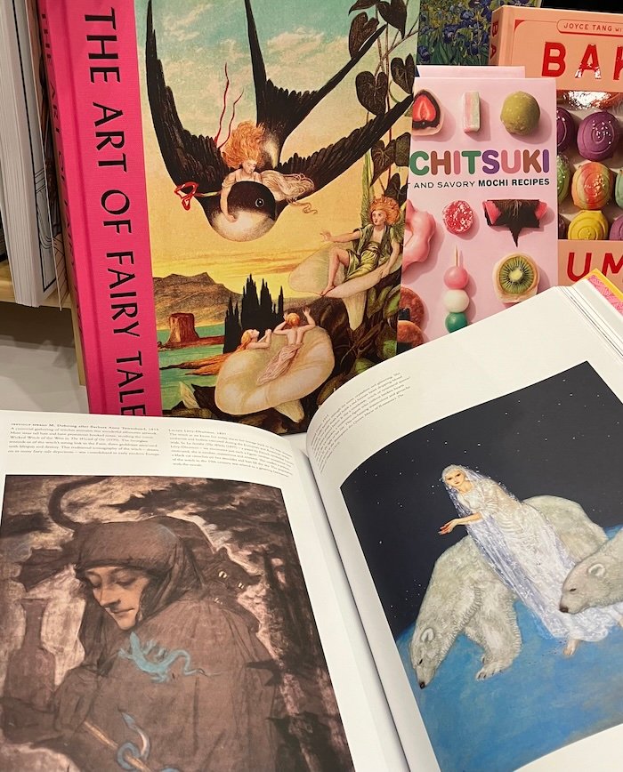 Book cover with image of small woman riding the back of a brown and white bird with black text running vertically along the wide bright pink spine Art of FairyTales. Open pages of the book in front with a picture of a woman wearing a long white dress surrounded by polar bears