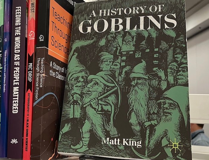 Book cover with black background and green image of small human figures wearing pointed hats and holding pick axes. Text in white reads A History of Goblins.